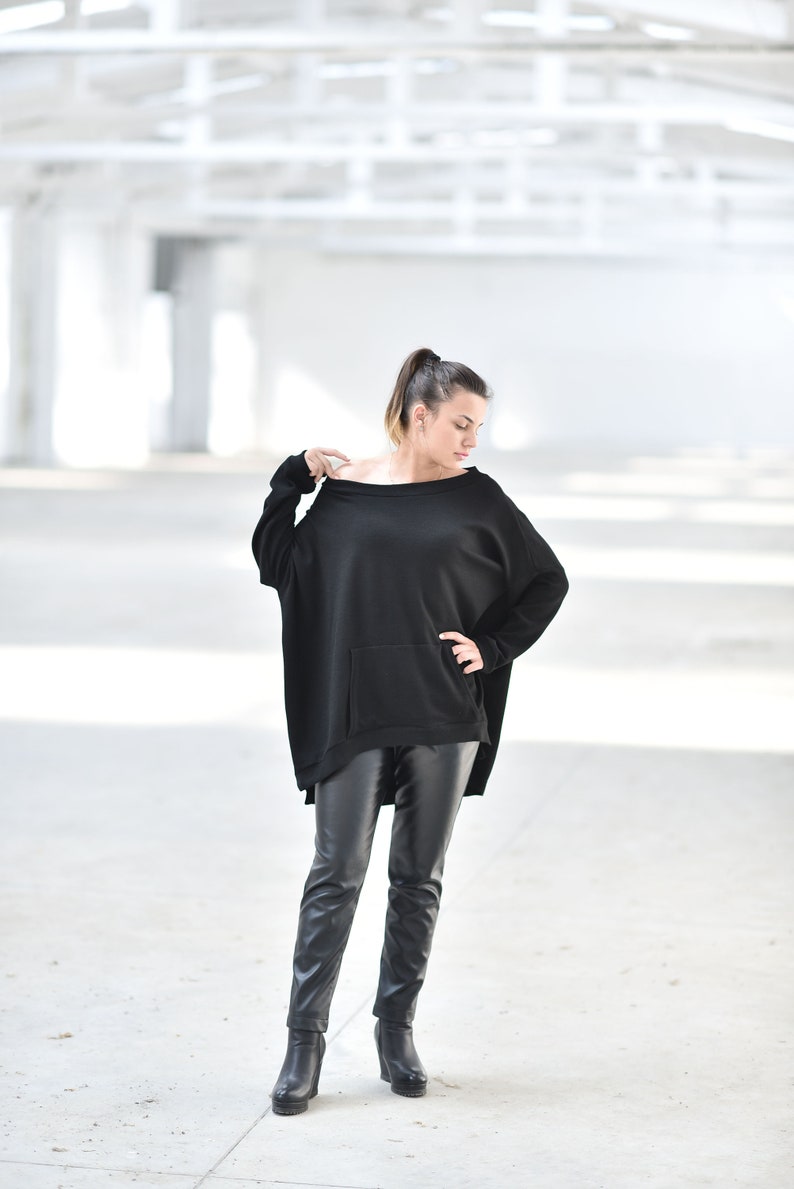 May include: A woman wearing a black oversized sweater with a pocket and black leather pants. She is standing in a white industrial setting.