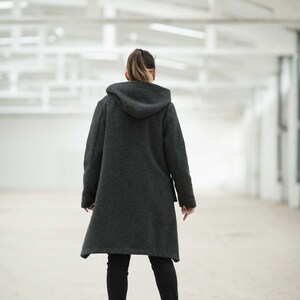 May include: A person wearing a dark gray hooded coat with a long hemline and black pants. The person is standing in a white, industrial-looking space.