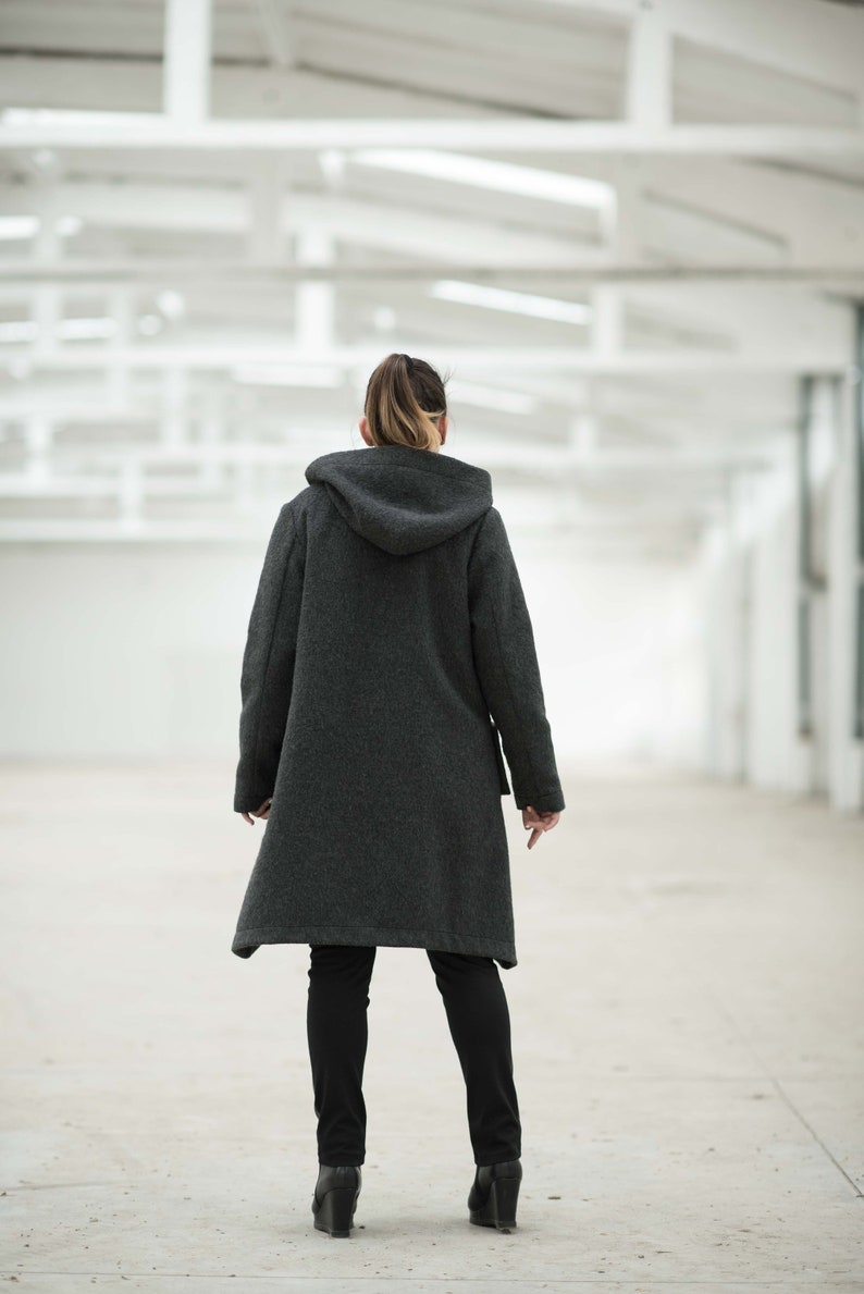 May include: A person wearing a dark gray hooded coat with a long hemline and black pants. The person is standing in a white, industrial-looking space.