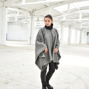 May include: A woman wearing a gray and black poncho with a zipper. She is standing in a white room with a concrete floor.