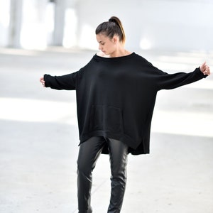 May include: A woman wearing a black oversized sweater with a pocket and black leather pants. She is wearing black ankle boots.