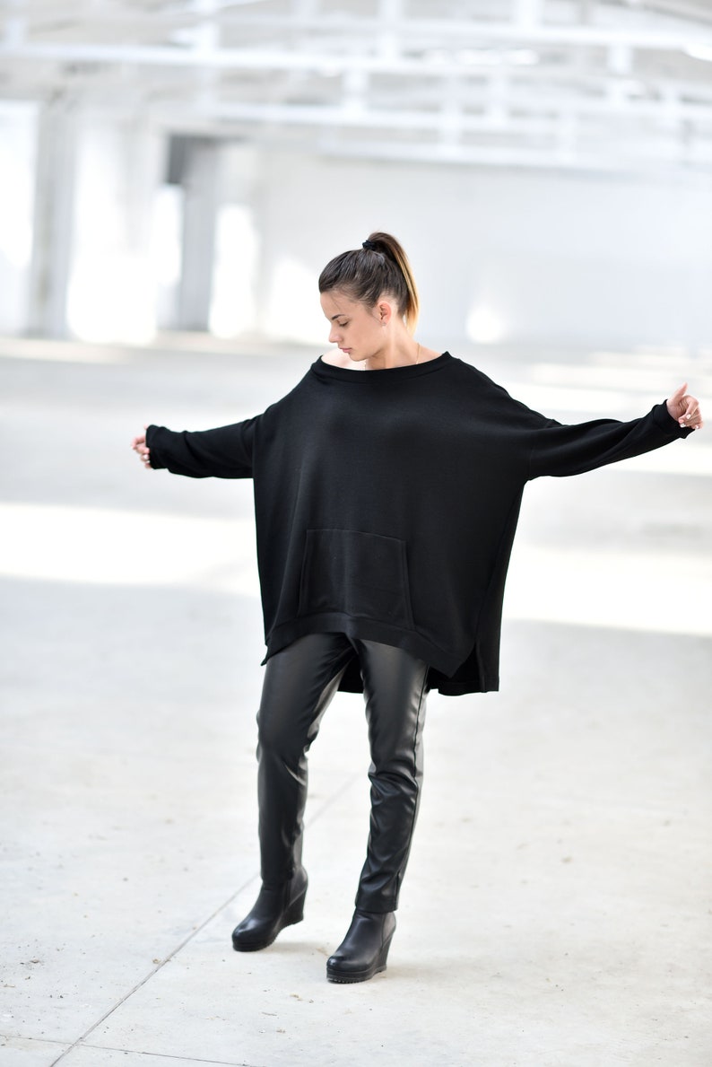 May include: A woman wearing a black oversized sweater with a pocket and black leather pants. She is wearing black ankle boots.