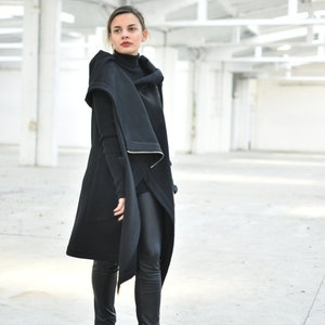 May include: A woman wearing a black outfit, including a long coat with a hood, a turtleneck, and black pants. The coat has a unique design with a draped front and a white trim. She is also wearing black boots.
