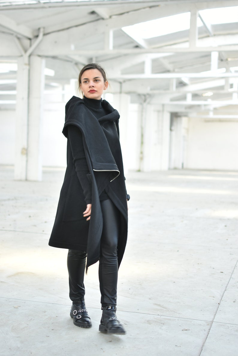 May include: A woman wearing a black outfit, including a long coat with a hood, a turtleneck, and black pants. The coat has a unique design with a draped front and a white trim. She is also wearing black boots.