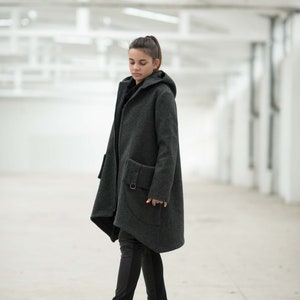 May include: A woman wearing a dark gray hooded coat with a long, asymmetrical hemline. The coat has a zipper closure and a large pocket on the left side. She is wearing black pants and black boots.