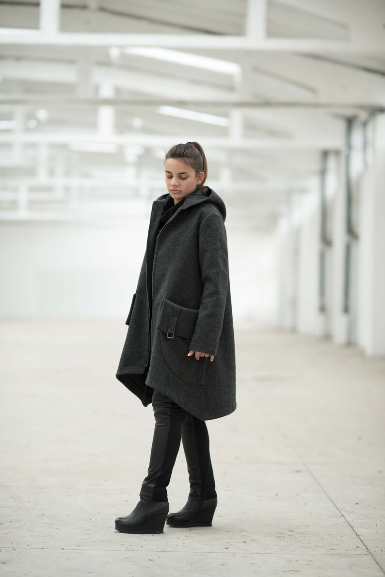 May include: A woman wearing a dark gray hooded coat with a long, asymmetrical hemline. The coat has a zipper closure and a large pocket on the left side. She is wearing black pants and black boots.