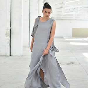 May include: A woman wearing a gray linen dress with a single sleeve and a bow detail at the waist. The dress has a high slit on one side and the woman is wearing white sneakers.
