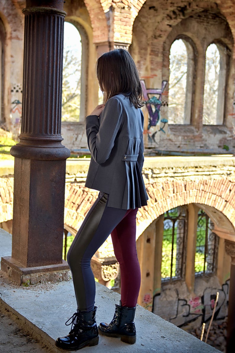 Puede incluir: Una mujer con un blazer gris con la espalda plisada, leggings negros y azules y leggings burdeos. Est&aacute; de pie en un edificio en ruinas con paredes de ladrillo y ventanas arqueadas.