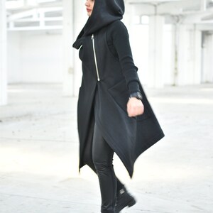 May include: A black hooded vest with a gold zipper, paired with black leggings and boots. The vest has a unique asymmetrical design. The outfit is completed with a black watch. The background is a white, open space.