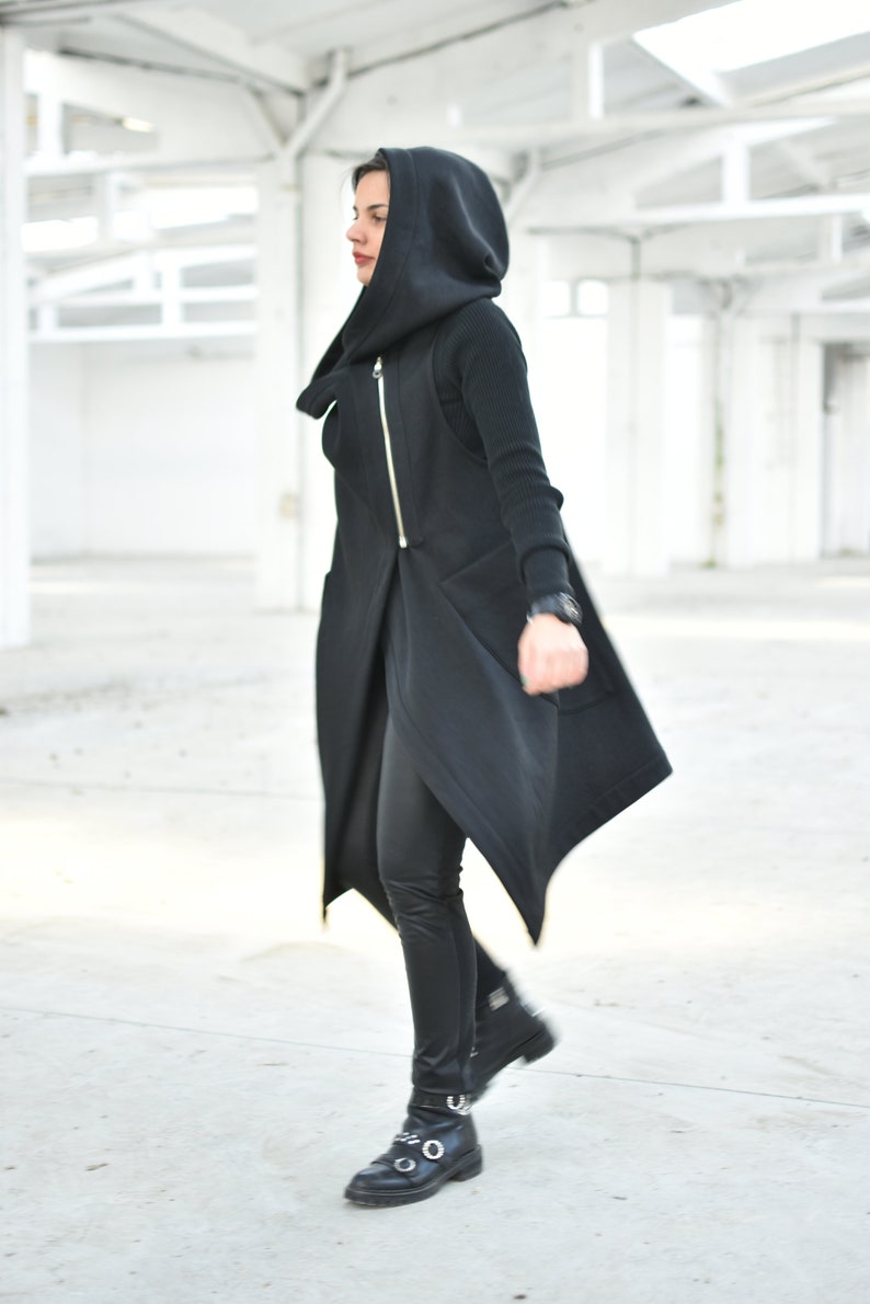 May include: A black hooded vest with a gold zipper, paired with black leggings and boots. The vest has a unique asymmetrical design. The outfit is completed with a black watch. The background is a white, open space.