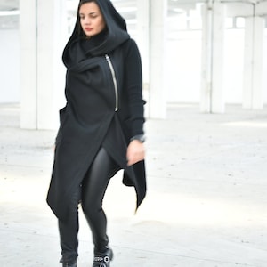 May include: A black hooded wrap coat with a diagonal zipper, paired with black leggings and black boots. The coat has a unique asymmetrical design. The boots have silver buckles.