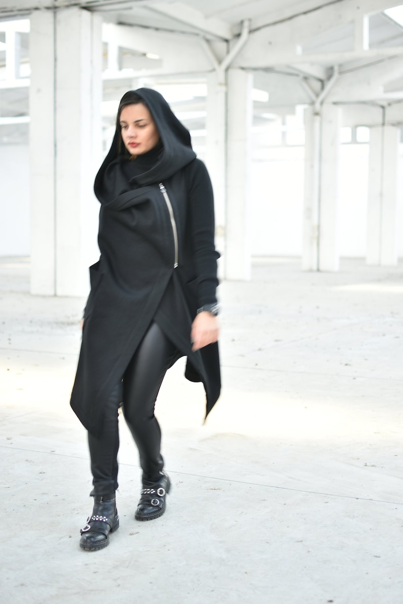 May include: A black hooded wrap coat with a diagonal zipper, paired with black leggings and black boots. The coat has a unique asymmetrical design. The boots have silver buckles.
