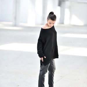 May include: A woman wearing a black oversized sweater with a long, asymmetrical hemline and black leather pants. She is wearing black wedge boots and standing in a white, industrial setting.
