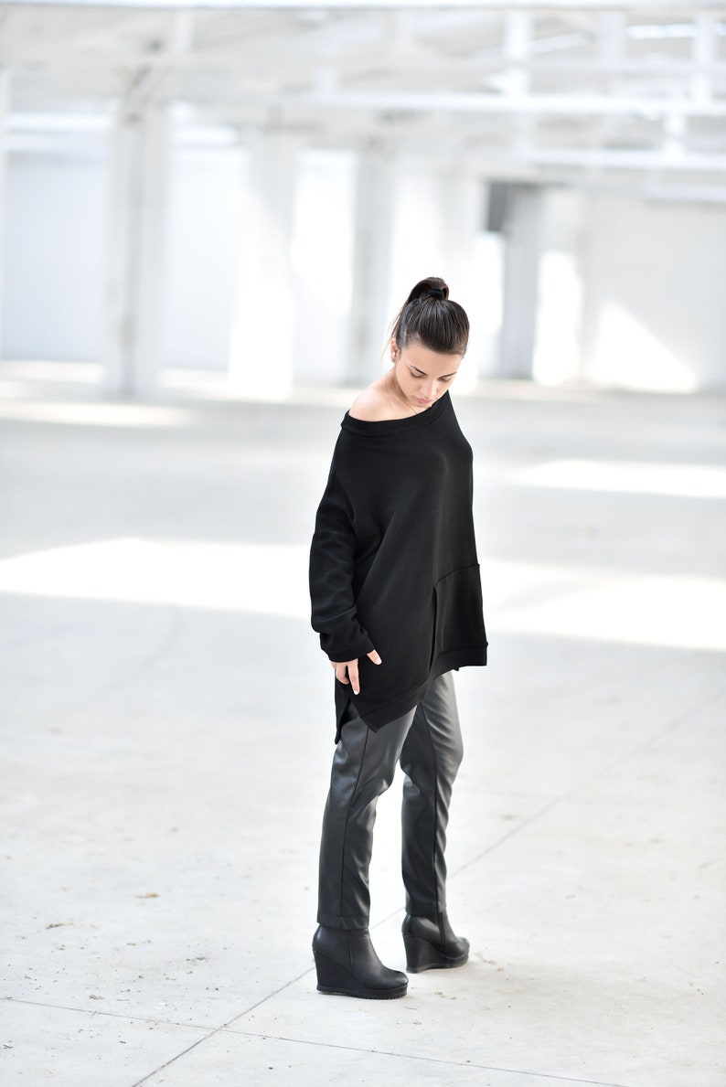 May include: A woman wearing a black oversized sweater with a long, asymmetrical hemline and black leather pants. She is wearing black wedge boots and standing in a white, industrial setting.
