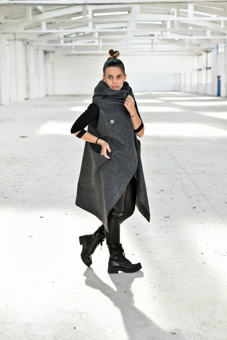May include: A woman wearing a long, gray, hooded vest with a high collar. She is wearing black pants and black boots. The vest has a unique asymmetrical design and a button closure.