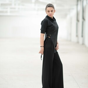May include: A woman wearing a black top with a side tie and black wide-leg trousers. She is standing in a white room with a white tiled floor.