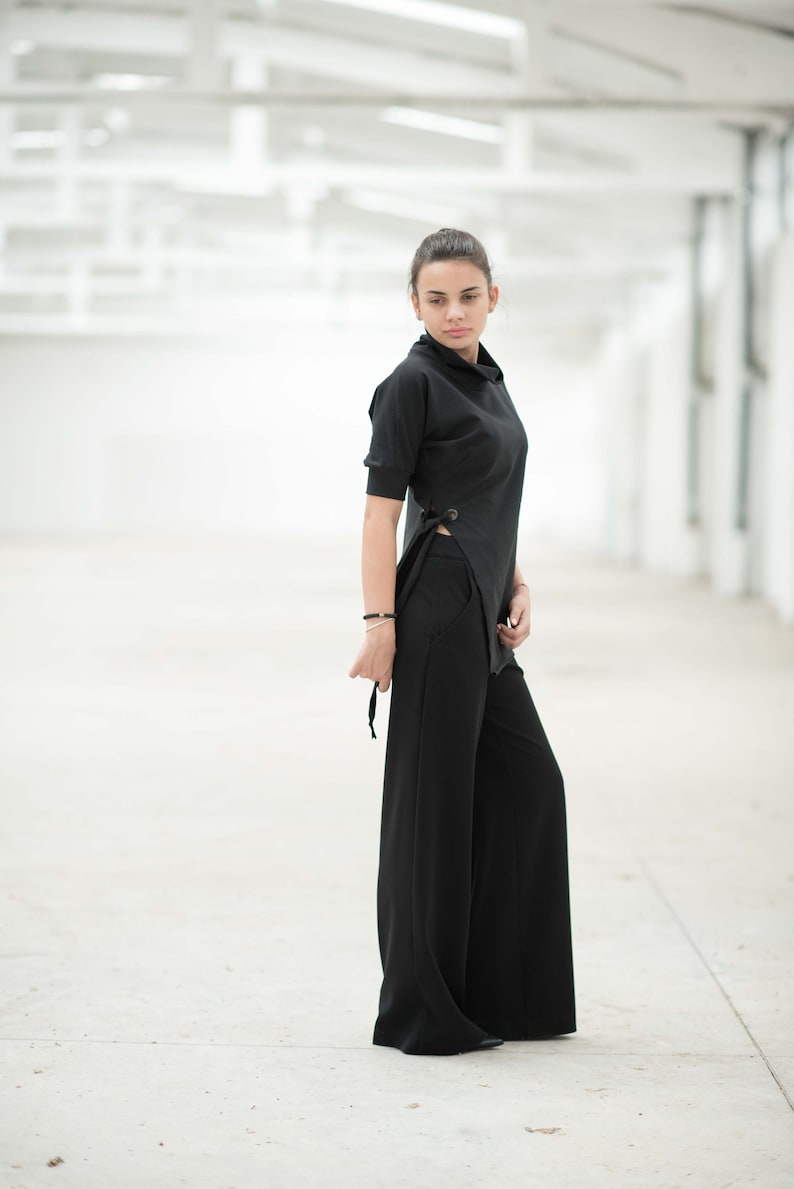 May include: A woman wearing a black top with a side tie and black wide-leg trousers. She is standing in a white room with a white tiled floor.