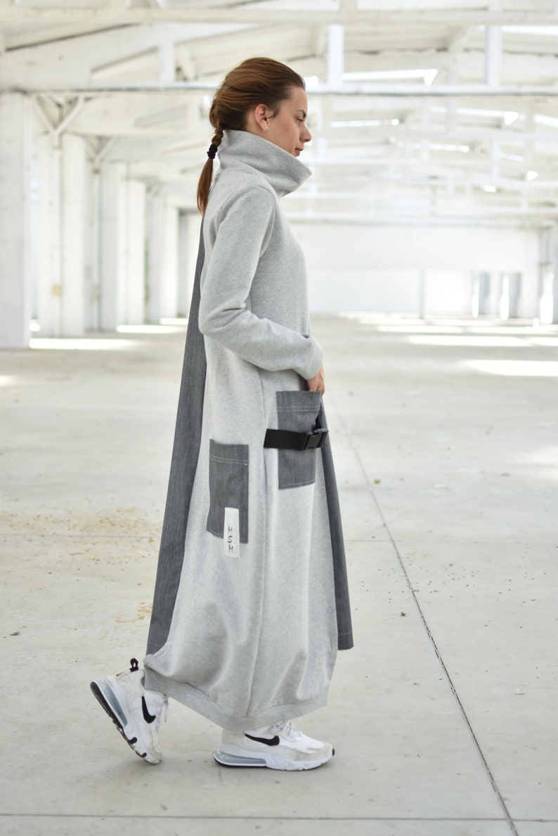 Puede incluir: Un vestido largo de manga larga gris y negro con cuello alto y cintur&oacute;n. El vestido tiene un bolsillo grande en la parte delantera y un bolsillo m&aacute;s peque&ntilde;o en el lateral. El vestido est&aacute; hecho de una tela suave y c&oacute;moda.