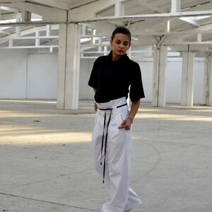 May include: A woman wearing a black short-sleeved shirt and white pants with a black belt. She is standing in a large, white, industrial space.