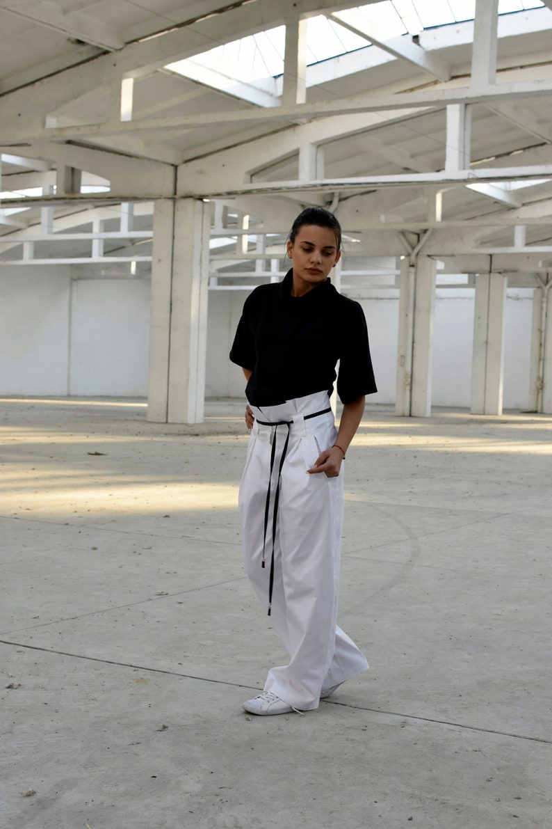 May include: A woman wearing a black short-sleeved shirt and white pants with a black belt. She is standing in a large, white, industrial space.