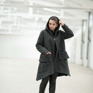 May include: A woman wearing a gray hooded coat with large pockets and black pants. She is standing in a white room with a concrete floor.