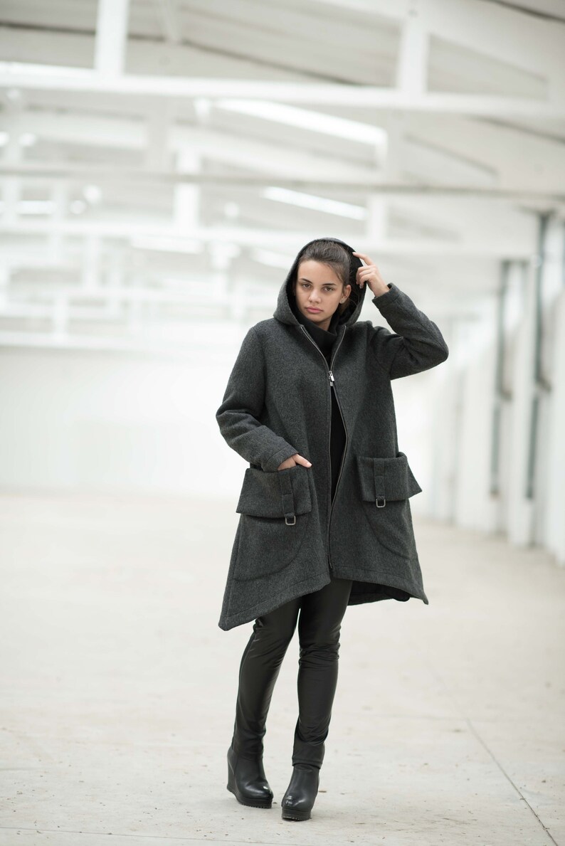 May include: A woman wearing a gray hooded coat with large pockets and black pants. She is standing in a white room with a concrete floor.