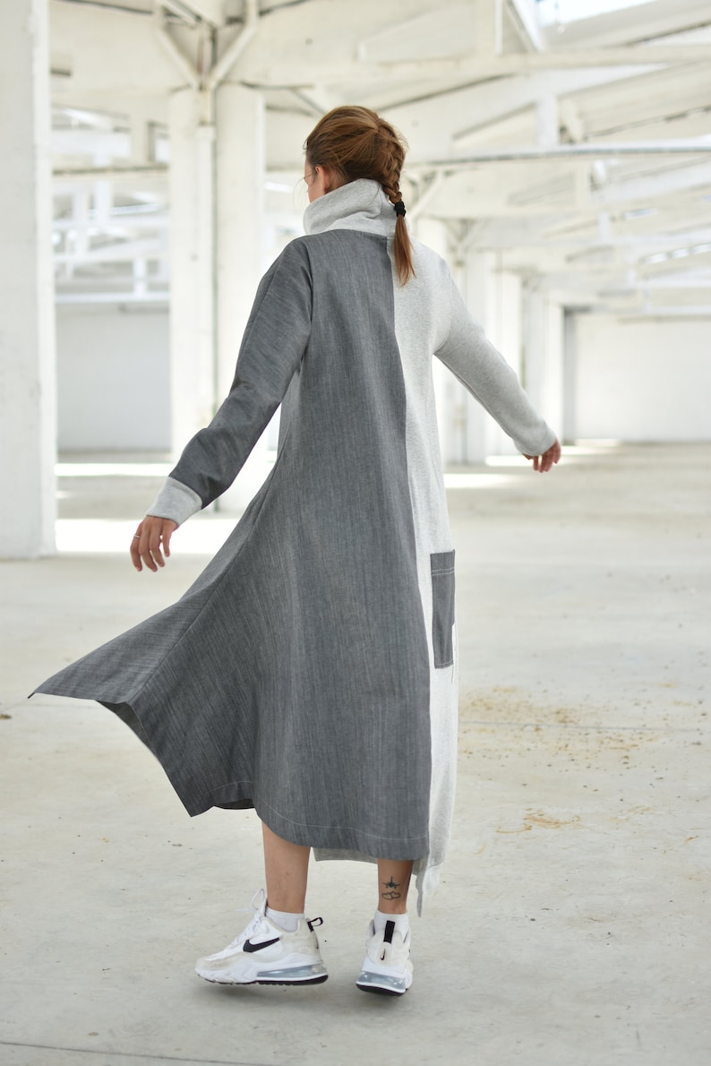 Puede incluir: Un vestido largo, gris y blanco con cuello alto y mangas largas. El vestido tiene un dobladillo asim&eacute;trico y fluido. La modelo lleva zapatillas blancas.