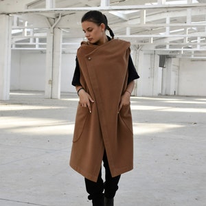 May include: A long brown sleeveless coat with a large collar and pockets. The coat is being worn by a person with dark hair and is standing in a white room.