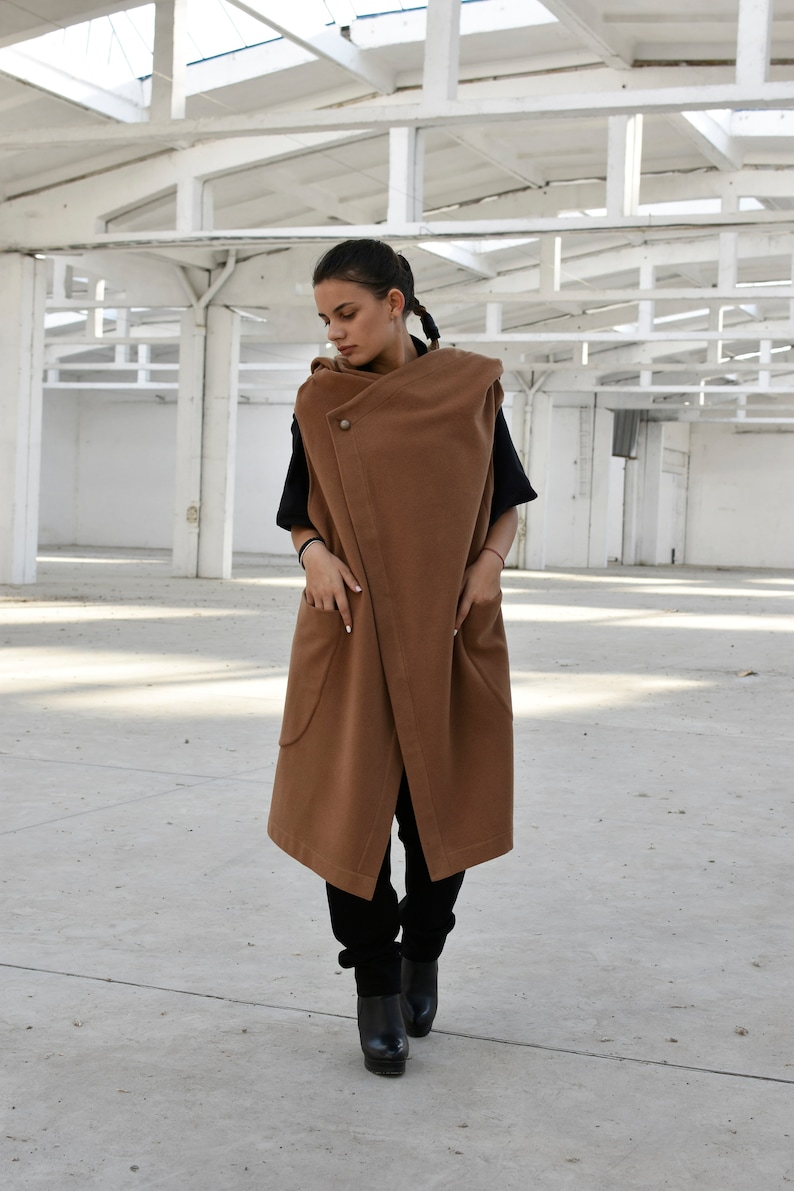 May include: A long brown sleeveless coat with a large collar and pockets. The coat is being worn by a person with dark hair and is standing in a white room.