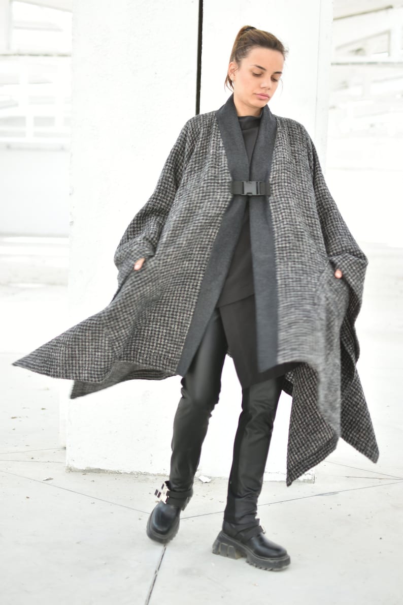 Pu&ograve; includere: Un cappotto a quadri bianco e nero con una cintura e fibbia nere. Il cappotto &egrave; indossato sopra un dolcevita nero e pantaloni neri. La persona indossa scarpe platform nere con fibbie.