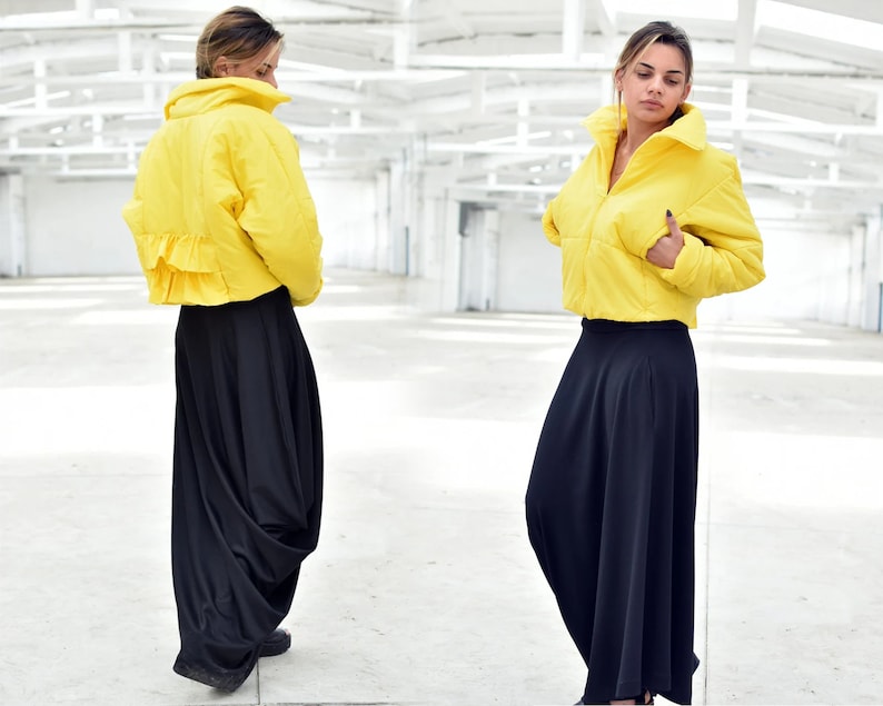 May include: A bright yellow cropped puffer jacket with a high collar and a ruffled hem detail, paired with a long, flowing black skirt. The jacket has a zipper closure and a puffy design. The outfit is set against a white background.