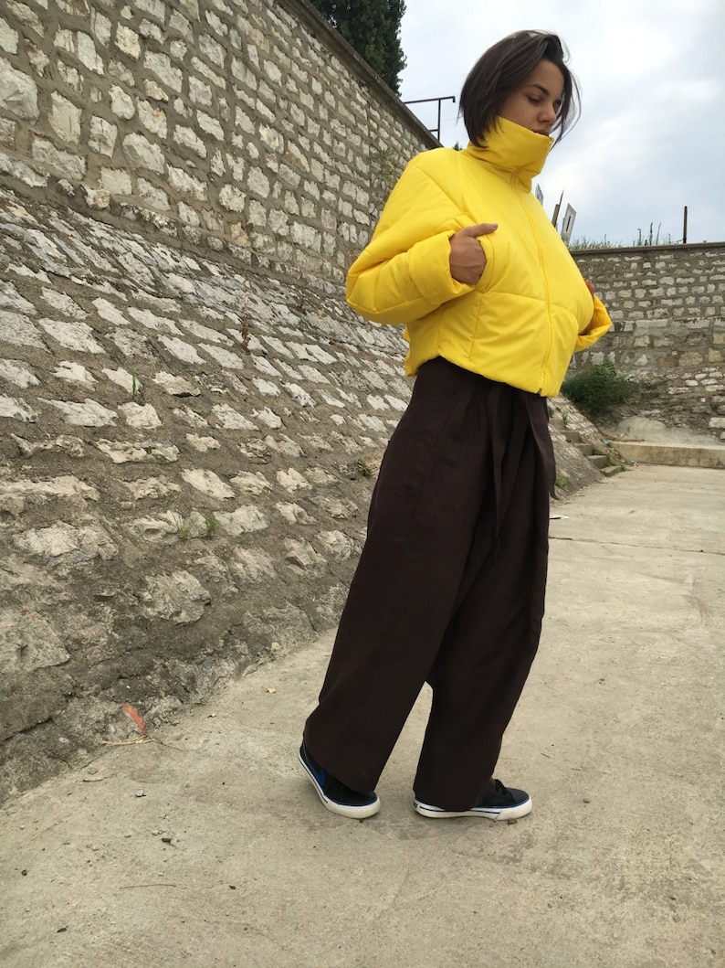 May include: A person wearing a bright yellow puffer jacket with a high collar and dark brown, wide-leg pants. The person is also wearing black sneakers with blue accents. The background features a stone wall and a cloudy sky.