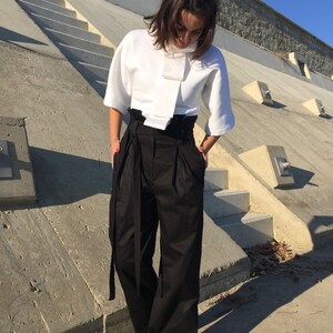 May include: A woman wearing a white, short-sleeved top with a large bow at the neck and black, high-waisted pants with a wide belt.