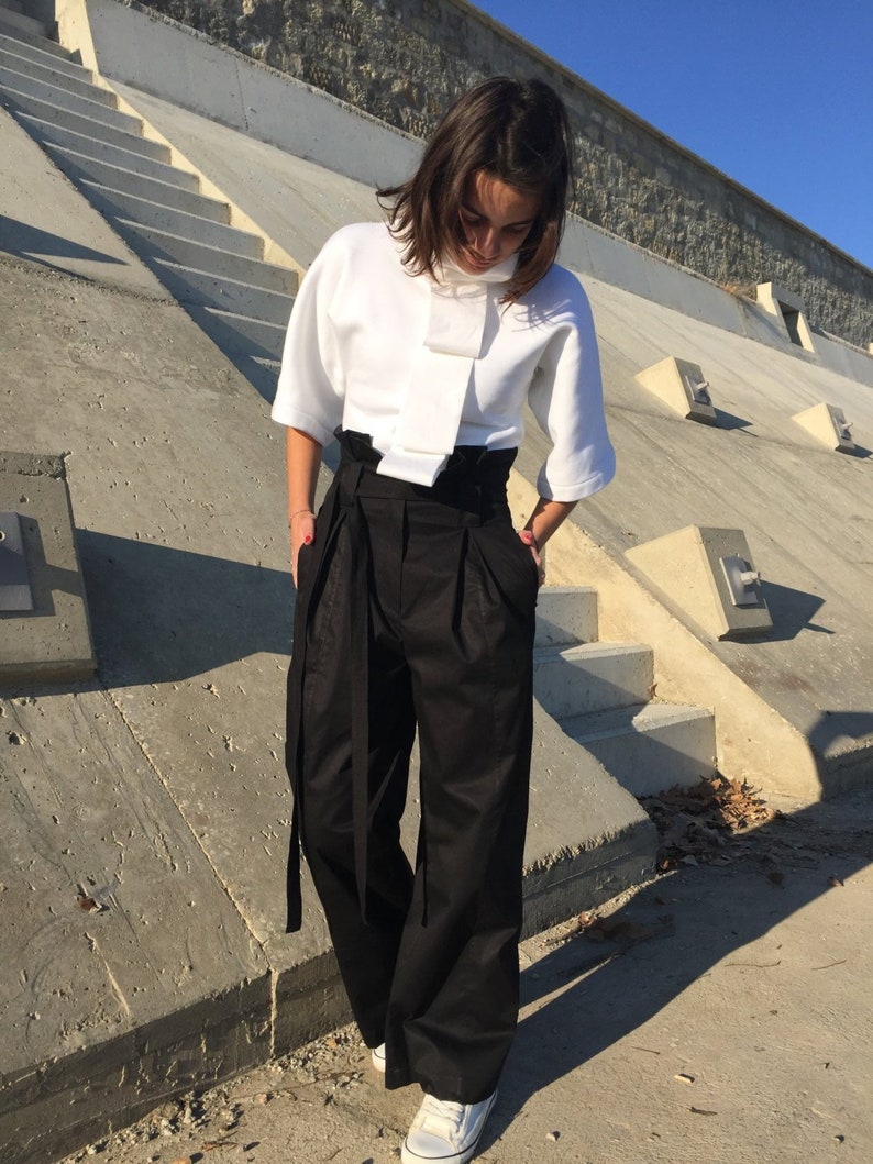 May include: A woman wearing a white, short-sleeved top with a large bow at the neck and black, high-waisted pants with a wide belt.