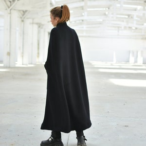 May include: A black long-sleeved cape with a high neck, worn by a person with long brown hair. The person is standing in a white industrial space.