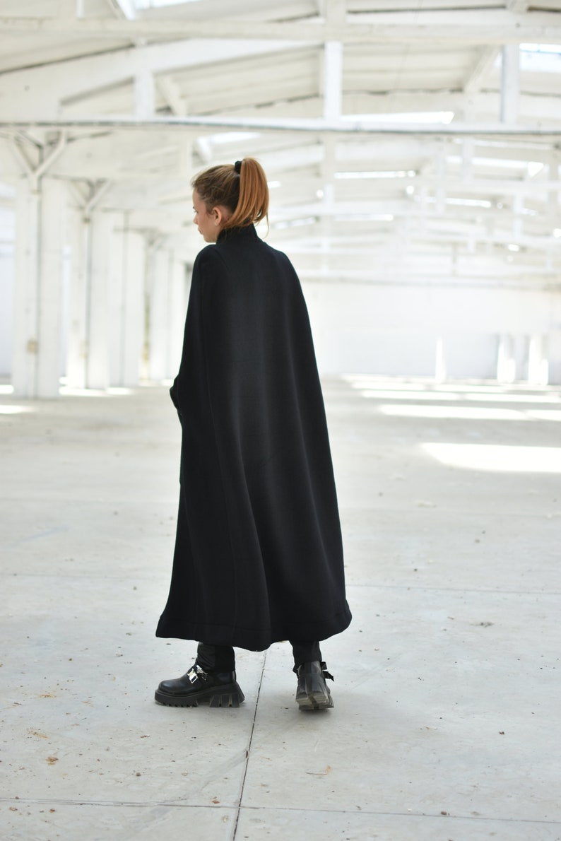 May include: A black long-sleeved cape with a high neck, worn by a person with long brown hair. The person is standing in a white industrial space.