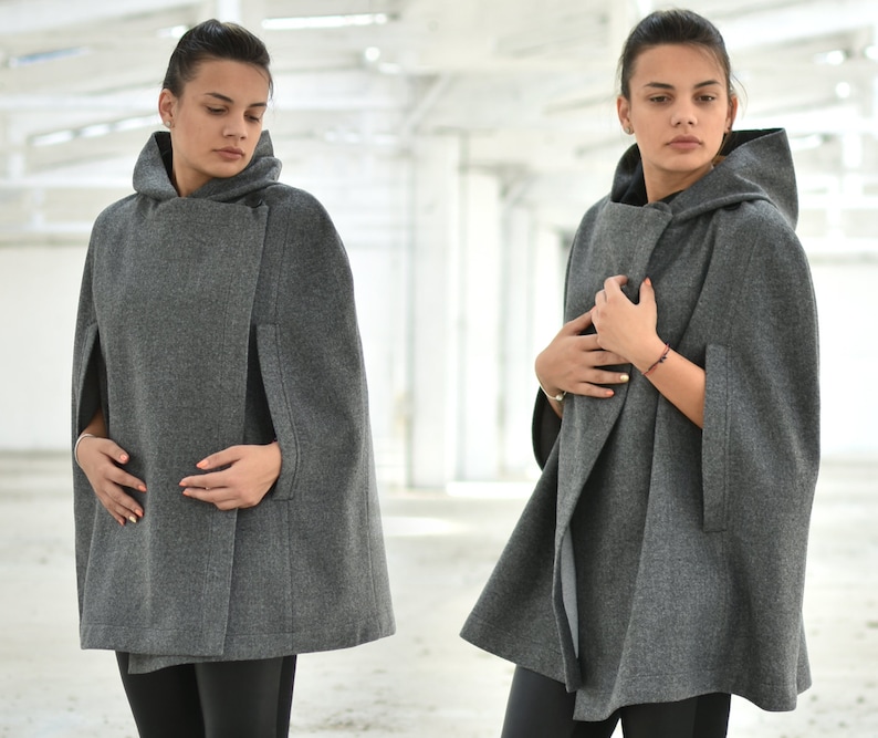 K&ouml;nnte beinhalten: Ein grauer Kapuzenumhang mit einem langen, flie&szlig;enden Design. Der Umhang hat einen Knopfverschluss und einen breiten Kragen.