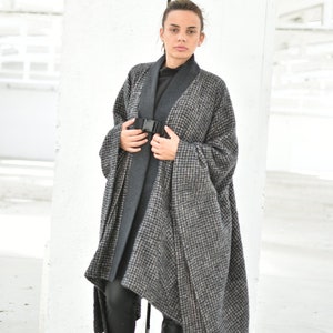 Pu&ograve; includere: Una donna indossa un lungo cappotto a quadri grigio e nero con una cintura nera. Indossa leggings neri e stivali neri.