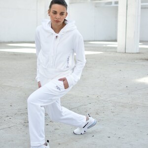 Puede incluir: Una sudadera con capucha blanca y pantalones de ch&aacute;ndal a juego con la palabra "HSH" bordada en la pierna izquierda. La modelo lleva zapatillas blancas con detalles negros.
