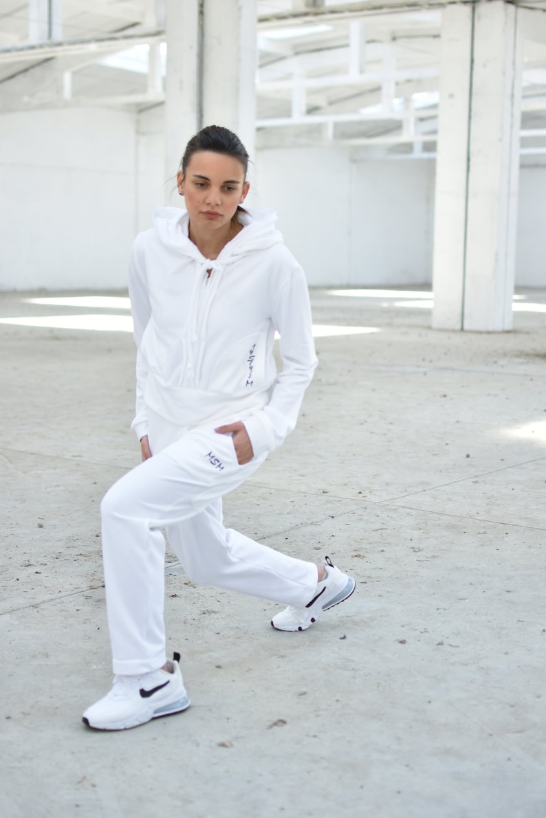Puede incluir: Una sudadera con capucha blanca y pantalones de ch&aacute;ndal a juego con la palabra "HSH" bordada en la pierna izquierda. La modelo lleva zapatillas blancas con detalles negros.