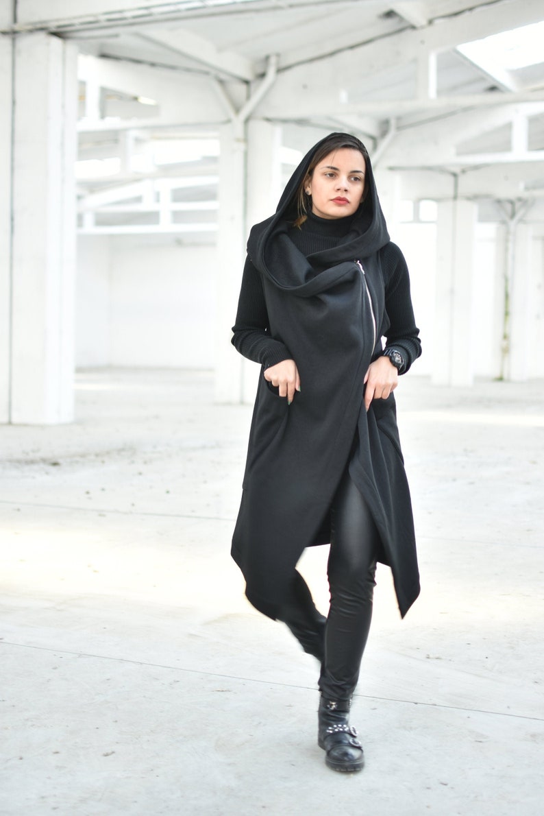 May include: A long, black hooded vest with a diagonal zipper closure. The vest is paired with a black turtleneck, black leggings, and black boots. The outfit is worn in a bright, open space with white pillars.