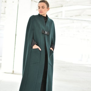 May include: A long, dark green cape with a black buckle and two pockets. The cape is made of a soft, warm fabric and has a relaxed fit.