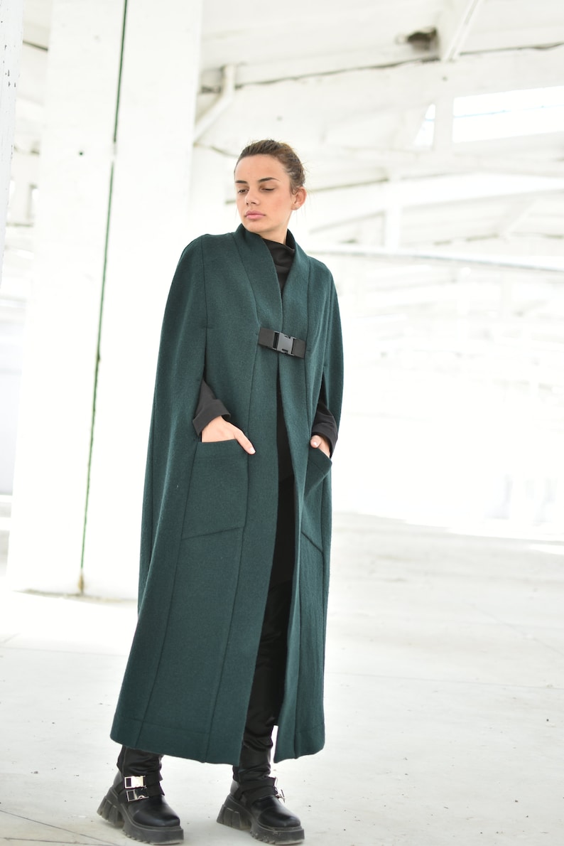 May include: A long, dark green cape with a black buckle and two pockets. The cape is made of a soft, warm fabric and has a relaxed fit.