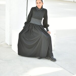 May include: A woman wearing a black and grey long-sleeved dress with a turtleneck. The dress has a gathered skirt and the woman is sitting on a white cube.