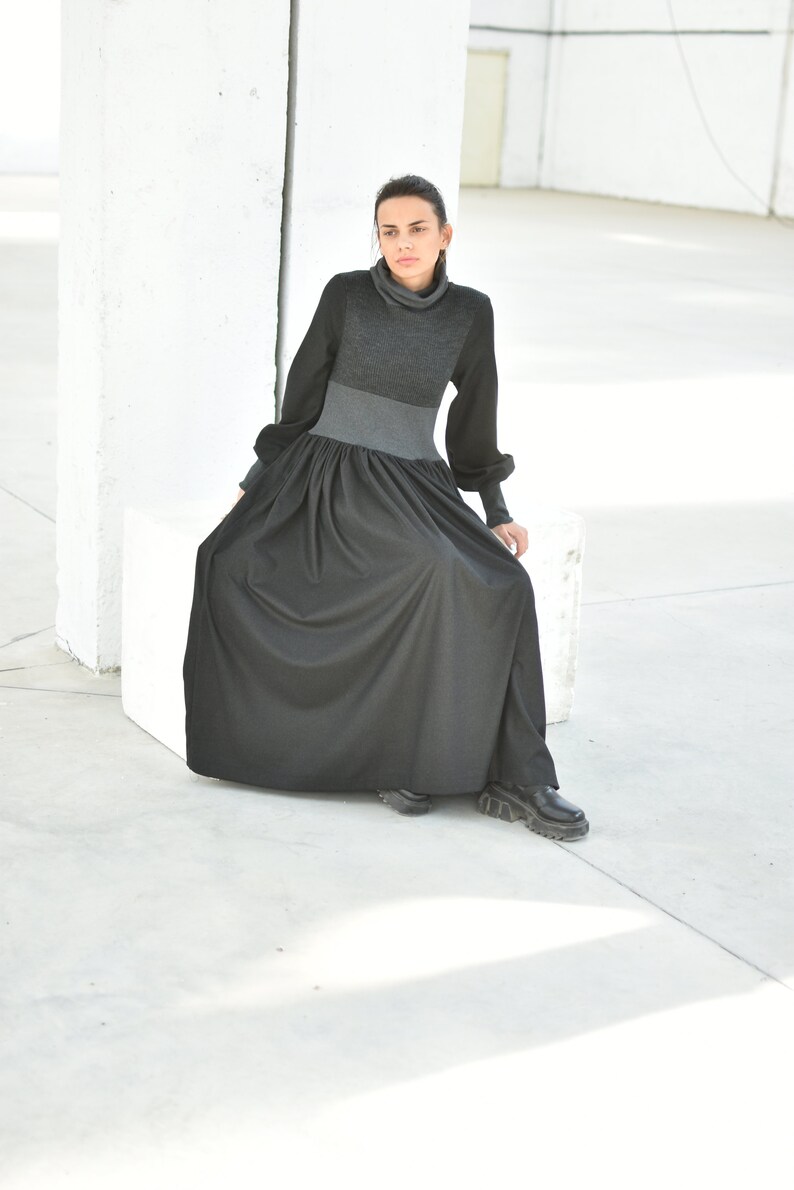 May include: A woman wearing a black and grey long-sleeved dress with a turtleneck. The dress has a gathered skirt and the woman is sitting on a white cube.