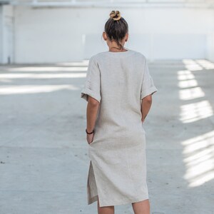 May include: A woman wearing a light beige linen dress with a side slit and short sleeves. She is standing in a large, empty, white room with a metal grid ceiling.