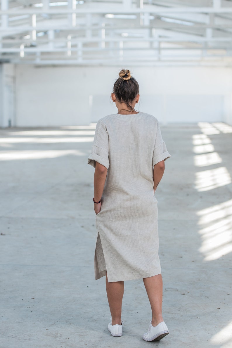 May include: A woman wearing a light beige linen dress with a side slit and short sleeves. She is standing in a large, empty, white room with a metal grid ceiling.