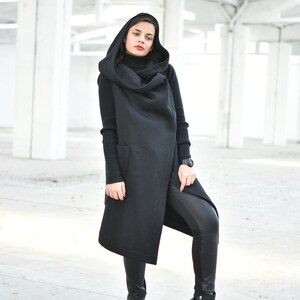 May include: A woman wearing a black hooded vest, black turtleneck, and black leather pants. The long vest has two pockets and a draped hood. She is also wearing black boots with silver accents. The background is a white industrial space.