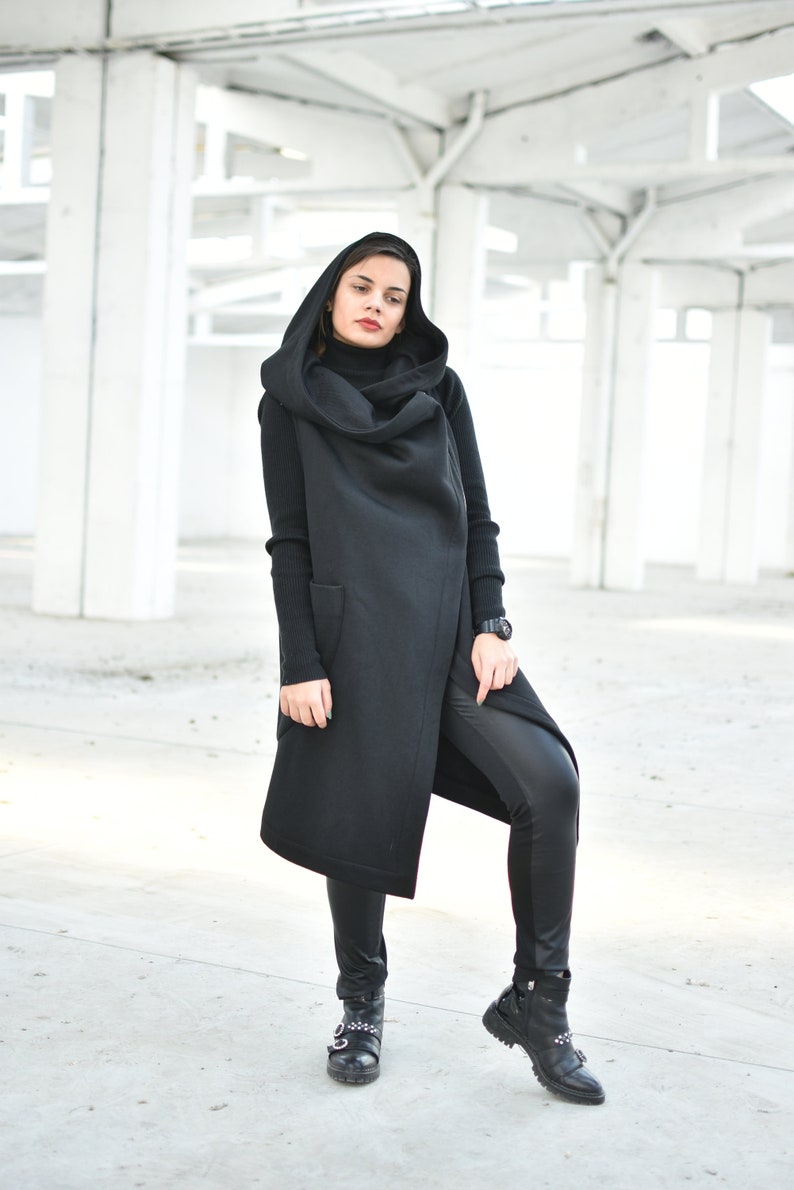 May include: A woman wearing a black hooded vest, black turtleneck, and black leather pants. The long vest has two pockets and a draped hood. She is also wearing black boots with silver accents. The background is a white industrial space.