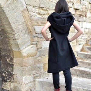 May include: A black sleeveless hooded vest with a long, flowing design. The vest has a large hood and a single pocket on the front. The person wearing the vest is standing on a set of stone steps.